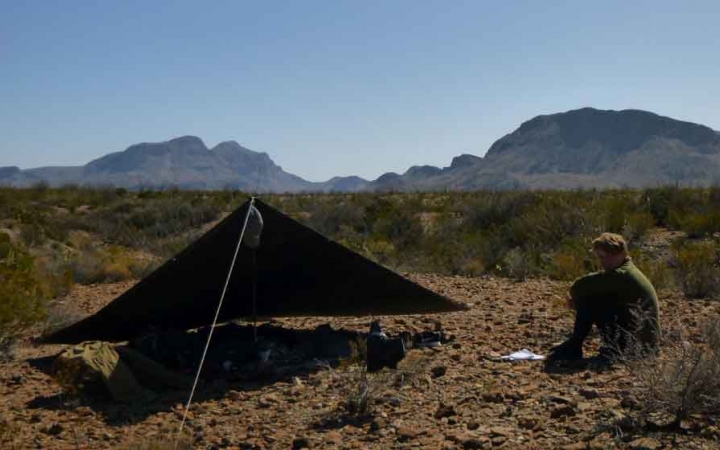 a person sits beside a tarp shelter in the desert with mountains in the background
